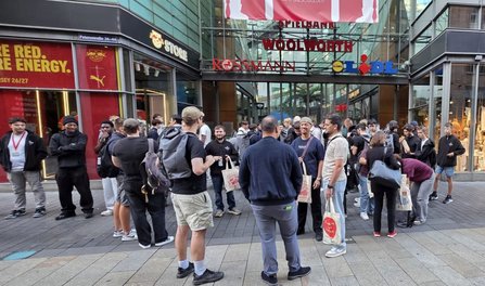Fußgängerbereich vor einem Einkaufszentrum mit den sichtbaren Schriftzügen „STORE“, „ROSSMANN“, „WOOLWORTH“ und „Lidl“. Mehrere Dutzend Personen stehen in Gruppen zusammen. Einige tragen helle Stofftaschen mit rotem Aufdruck. Im Vordergrund stehen Personen mit Rucksäcken und Taschen, im Hintergrund weitere Personen vor Schaufenstern und Eingängen.