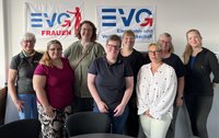 Acht Personen stehen in einem Innenraum vor zwei aufgehängten Bannern. Auf dem linken Banner steht „EVG FRAUEN“, auf dem rechten „EVG Eisenbahn- und Verkehrsgewerkschaft“. Die Personen sind in zwei Reihen angeordnet und blicken zur Kamera. Im Vordergrund stehen teilweise sichtbare schwarze Stühle und ein runder Tisch. Die Wände sind hell, das Licht fällt von links in den Raum.