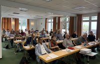 Seminarraum mit mehreren Reihen von Tischen. Rund dreißig Personen sitzen auf Stühlen und blicken nach vorn. Auf den Tischen liegen Unterlagen, Notizblätter, Getränke und Gläser. Im Vordergrund steht ein Beamer auf einem kleinen Tisch. Der Raum hat große Fenster mit Vorhängen, helle Wände, Deckenbeleuchtung und einen Ausgang im Hintergrund.