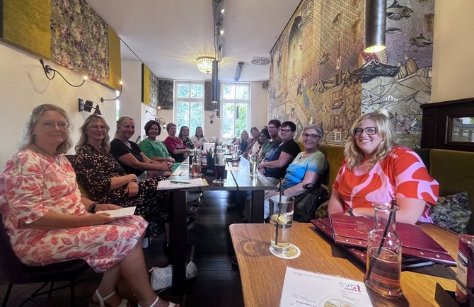 Mehrere Personen sitzen an zwei langen Tischen in einem Restaurant oder Café. Auf den Tischen stehen Gläser, Karaffen, Speisekarten und Unterlagen. An den Wänden hängen großformatige Kunstwerke, und im Hintergrund befinden sich hohe Fenster.