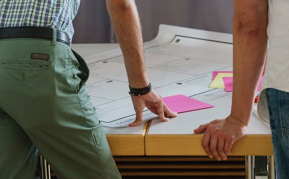 Zwei Personen lehnen sich mit den Händen über einen zusammengestellten Tisch. Auf dem Tisch liegen große bedruckte Papierbögen und mehrere pinkfarbene Haftnotizen. Sichtbar sind Arme, Hände sowie Teile der Kleidung; im Hintergrund befindet sich eine unscharfe Wandfläche.