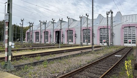 Mehrgleisiger Bahnbereich vor einer großen Halle mit mehreren hohen Toren. Oberleitungen, Masten und Gleise verlaufen im Vordergrund. Die Hallenfassade ist grau mit hellvioletten Torumrandungen.