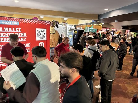 Innenraum mit Informationsständen und vielen Besuchenden. Links steht ein großer roter Aufsteller mit der Aufschrift „EVG JUGEND NRW“, einem QR-Code und teilweise sichtbarem Text „DU WILLST NICHTS MEHR VERPASSEN?“. Vor dem Aufsteller stehen mehrere Personen. Im Hintergrund sind weitere Stände mit den Beschriftungen „DEVK“ und „BAHN BKK“ sowie Automaten und eine große dekorative Figur zu sehen.