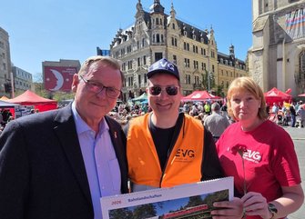 Drei Personen stehen im Vordergrund eines belebten Platzes im Freien. Die mittlere Person trägt eine orangefarbene Warnweste und eine dunkle Kappe mit weißer Aufschrift sowie eine Sonnenbrille und hält einen Kalender mit der Aufschrift „2026 Bahnlandschaften“, auf dem ein roter Zug in einer Landschaft abgebildet ist. Die Person links trägt ein dunkles Jackett und ein helles Hemd, die Person rechts ein rotes T‑Shirt und hält eine einzelne rote Blume. Im Hintergrund sind zahlreiche Menschen, rote Zelte, Fahnen sowie mehrstöckige historische Gebäude und eine Kirche zu sehen.