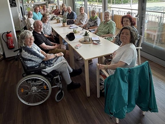 Mehrere Personen sitzen gemeinsam an einem langen Tisch in einem hellen Raum und schauen in die Kamera. Insgesamt sind rund ein Dutzend Personen an zwei zusammengestellten Tischen platziert, die längs durch den Raum verlaufen. Die Personen sitzen dicht beieinander auf Stühlen; eine Person im Vordergrund links sitzt in einem Rollstuhl. Auf den Tischen stehen kleine Pflanzen, Tassen, Unterlagen und einige persönliche Gegenstände. Der Raum ist hell und hat große Fenster auf der rechten Seite, durch die viel Tageslicht einfällt. Durch die Fenster ist ein Balkon oder Außenbereich mit Geländer und Pflanzen sichtbar. Der Boden ist dunkel und glatt, vermutlich Holz oder Laminat. Im linken hinteren Bereich stehen weitere Pflanzen sowie ein roter Feuerlöscher an der Wand. Die Personen sind zum Teil einander zugewandt, viele blicken direkt in die Kamera. Die Gruppe wirkt wie bei einem Treffen oder einer gemütlichen Runde an einem Tisch, möglicherweise bei Kaffee oder einer Besprechung.