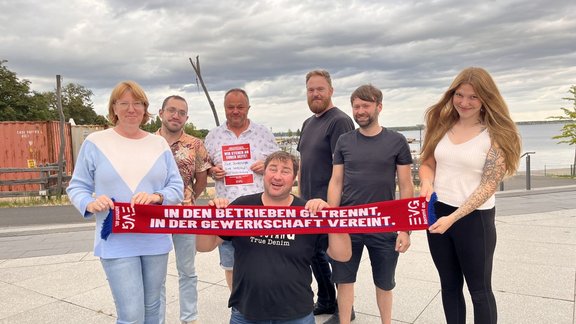 Sieben Personen stehen auf einer Terrasse am Wasser unter einem bewölkten Himmel. Eine Person kniet vorne und hält gemeinsam mit zwei Personen einen roten Schal mit der Aufschrift „IN DEN BETRIEBEN GETRENNT, IN DER GEWERKSCHAFT VEREINT.“. In der Mitte hält eine Person ein rotes Schild mit dem Text „WIR STEHEN AN EURER SEITE!“ und einem EVG-Logo. Im Hintergrund sind ein Gewässer, Geländer, Bäume und ein Container zu sehen.
