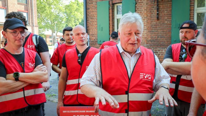 Mehrere Personen in roten EVG-Westen stehen vor einer Backsteinfassade. Eine Person im Vordergrund spricht mit erhobenen Händen zu einer Gruppe. Im unteren Bildbereich ist ein rotes Schild mit der Überschrift „MERKT EUCH DAS:“ teilweise sichtbar.