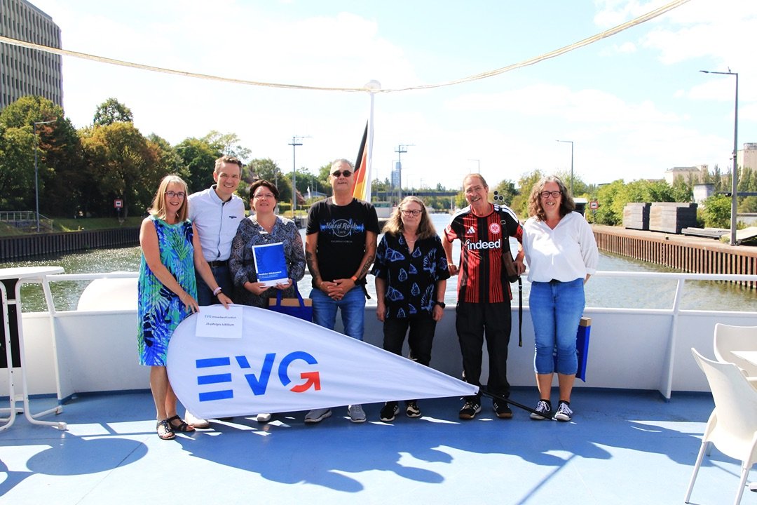 Sieben Personen stehen auf dem Außendeck eines Schiffes hinter einer großen EVG-Fahne und posieren für ein Gruppenfoto. Die Personen stehen nebeneinander auf einem hellblauen Schiffsdeck. Vor der Gruppe liegt eine weiße Fahne mit dem blauen und roten EVG-Logo. Eine Person hält eine blaue Mappe in den Händen. Im Hintergrund ist ein Mast mit einer Deutschlandflagge zu sehen. Auf dem Deck stehen außerdem weiße Tische und Stühle.