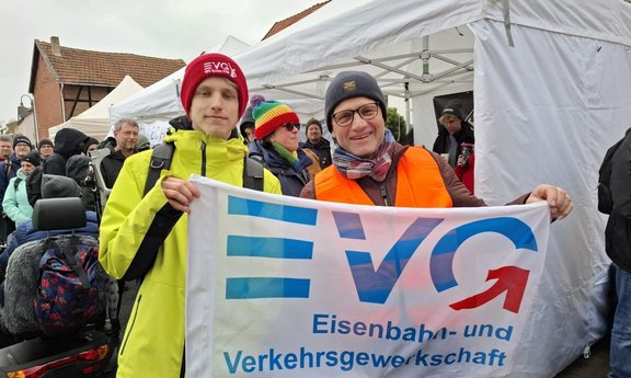 Zwei Personen stehen im Vordergrund dicht nebeneinander und halten ein Banner mit dem Logo und der Aufschrift „EVG Eisenbahn- und Verkehrsgewerkschaft“. Die linke Person trägt eine helle Jacke und eine rote Mütze mit EVG-Schriftzug. Die rechte Person trägt eine orangefarbene Warnweste, eine dunkle Mütze und einen Schal. Im Hintergrund sind mehrere weitere Personen, ein weißes Pavillonzelt und Häuser zu sehen.