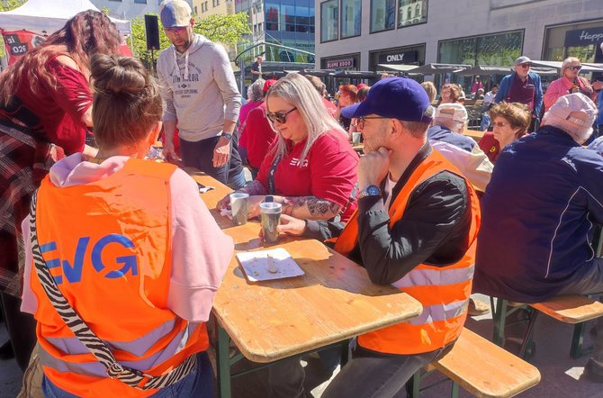 Mehrere Personen sitzen an einer Holztischgarnitur auf einem Platz. Zwei Personen tragen orangefarbene Warnwesten mit der Aufschrift „EVG“. Auf dem Tisch stehen Becher und ein Teller mit Essensresten. Um den Tisch herum stehen weitere Personen, im Hintergrund sind viele Menschen, Stände und Gebäude zu sehen.
