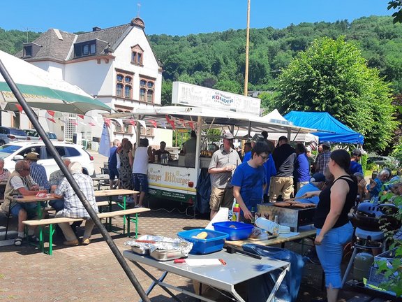 Ein belebter Außenbereich zeigt einen Getränkestand und einen Grill mit mehreren Personen, die essen, trinken oder sich unterhalten. Das Bild zeigt eine Szene im Freien bei sonnigem Wetter. In der Mitte steht ein weißer Getränkewagen mit Zapfanlage, über dem ein Schild mit der Aufschrift „Getränke Kondor“ zu sehen ist. Mehrere Personen stehen davor und holen Getränke oder sprechen miteinander. Rechts im Vordergrund befindet sich ein Grillstand: Auf einem Grill liegen mehrere Würstchen, daneben stehen Schalen, Besteck und Flaschen mit Saucen. Eine Person bedient den Grill, während eine andere Person daneben steht.