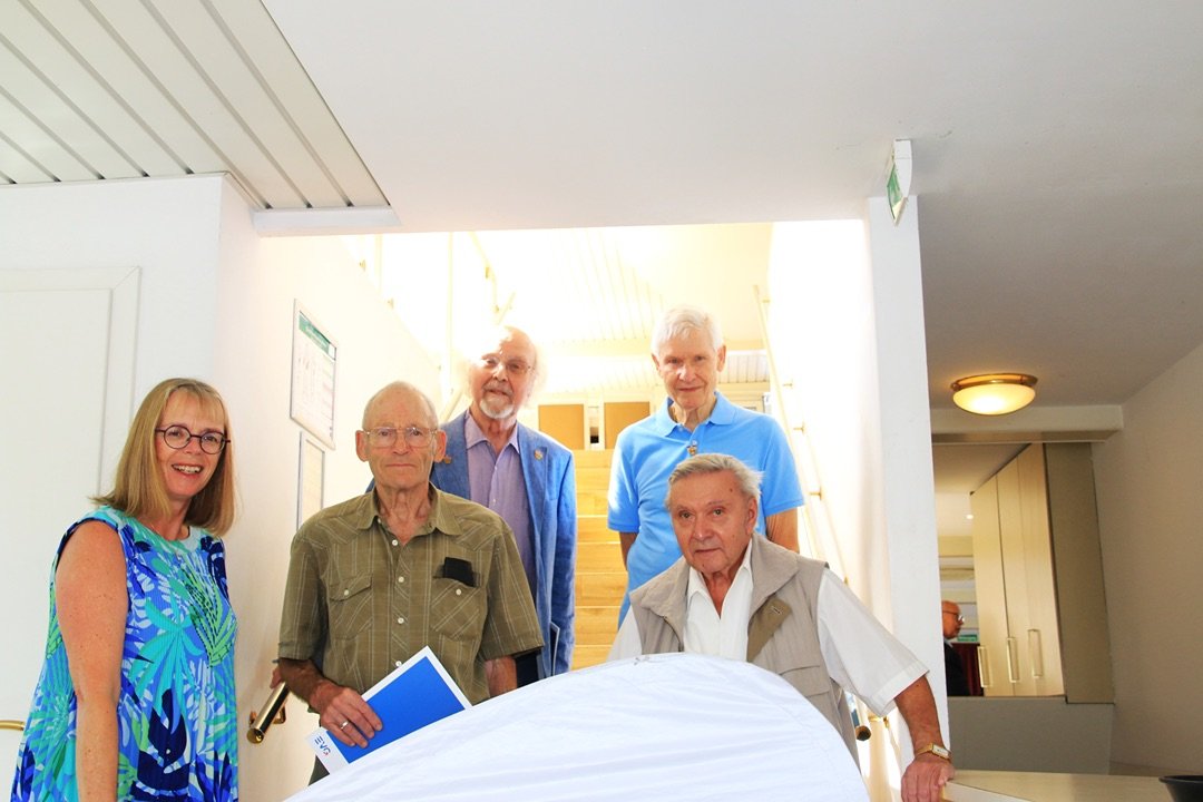 Fünf Personen stehen im Innenbereich eines Schiffes an einem Treppenaufgang und posieren für ein Gruppenfoto. Die Gruppe befindet sich vor einer hell beleuchteten Treppe mit Handläufen. Eine Person hält eine blaue Mappe in der Hand. Im Vordergrund ist teilweise eine weiße EVG-Fahne zu sehen. Die Personen stehen auf unterschiedlichen Ebenen, einige vor der Treppe, andere auf den Stufen dahinter.