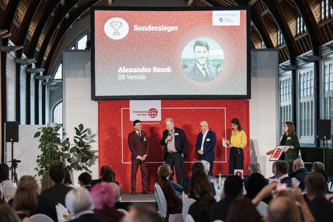 In einer großen Halle mit Holzdach und hohen Fenstern steht eine Bühne vor sitzendem Publikum. Auf der Bühne stehen mehrere Personen nebeneinander. Hinter ihnen ist eine große Leinwand mit rotem Hintergrund zu sehen. Auf der Leinwand steht gut lesbar „Sondersieger“ sowie der Name „Alexander Rezek“ und darunter „DB Vertrieb“. Weitere grafische Elemente und Logos sind sichtbar. Pflanzen, Mikrofone und ein Rednerpult befinden sich auf der Bühne.