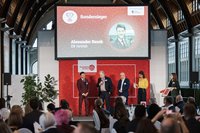 In einer großen Halle mit Holzdach und hohen Fenstern steht eine Bühne vor sitzendem Publikum. Auf der Bühne stehen mehrere Personen nebeneinander. Hinter ihnen ist eine große Leinwand mit rotem Hintergrund zu sehen. Auf der Leinwand steht gut lesbar „Sondersieger“ sowie der Name „Alexander Rezek“ und darunter „DB Vertrieb“. Weitere grafische Elemente und Logos sind sichtbar. Pflanzen, Mikrofone und ein Rednerpult befinden sich auf der Bühne.