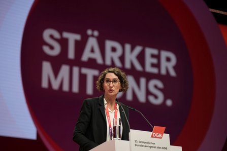 Person an einem Redepult auf einer Bühne. Im Hintergrund ein großes rundes rotes Motiv mit dem weißen Text „STÄRKER MIT UNS.“. Auf dem Pult steht ein Schild mit der Aufschrift „DGB“ und „23. Ordentlicher Bundeskongress des DGB“. Auf dem Pult stehen ein Mikrofon und ein Glas Wasser.
