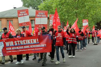 Demonstrationszug auf einer Straße vor Backsteingebäuden und Bäumen. Zahlreiche Teilnehmende tragen rote EVG-Fahnen sowie rote Westen und T‑Shirts. Im Vordergrund wird ein großes rotes Banner getragen mit der Aufschrift: „WIR STREIKEN! KEIN ABSCHLUSS OHNE GEWERKSCHAFT @GBM: Unsere Forderungen sind kein Hexenwerk!“. Mehrere Schilder mit der Überschrift „MERKT EUCH DAS:“ werden hochgehalten.