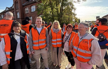 Mehrere Personen stehen auf einem gepflasterten Platz im Freien und tragen orangefarbene Warnwesten mit EVG-Logo und reflektierenden Streifen. Im Vordergrund stehen fünf Personen nebeneinander und blicken zur Kamera. Im Hintergrund befinden sich weitere Personen in orangefarbenen Westen, ein Backsteingebäude mit Fenstern, Bäume und eine Straße. Das Bild ist bei hellem Sonnenlicht aufgenommen, mit Schatten auf dem Boden und Gegenlicht von rechts. Sichtbarer Text auf einigen Westen: „EVG“ sowie „GEMEINSAM GEHT MEHR“.