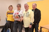 Vier Personen stehen nebeneinander in einem Innenraum vor einer hellen Wand mit einem großen gelben Wandpaneel. Die zweite Person von links hält einen dekorierten Geschenkkorb mit Schleife und einem kleinen Holzhaus. Links steht eine Person in einem gelben Pullover mit mehrfarbigen Streifen, daneben eine Person in weißem Oberteil und dunkler Hose. Rechts davon steht eine Person in einem hellgelben Hemd mit aufgedrucktem Text, daneben eine Person in dunkler Jacke und dunkler Hose. Im Vordergrund sind die Rückenlehne eines Stuhls und ein Kleidungsstück zu sehen, rechts ein Teil eines Tisches.