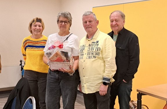 Vier Personen stehen nebeneinander in einem Innenraum vor einer hellen Wand mit einem großen gelben Wandpaneel. Die zweite Person von links hält einen dekorierten Geschenkkorb mit Schleife und einem kleinen Holzhaus. Links steht eine Person in einem gelben Pullover mit mehrfarbigen Streifen, daneben eine Person in weißem Oberteil und dunkler Hose. Rechts davon steht eine Person in einem hellgelben Hemd mit aufgedrucktem Text, daneben eine Person in dunkler Jacke und dunkler Hose. Im Vordergrund sind die Rückenlehne eines Stuhls und ein Kleidungsstück zu sehen, rechts ein Teil eines Tisches.