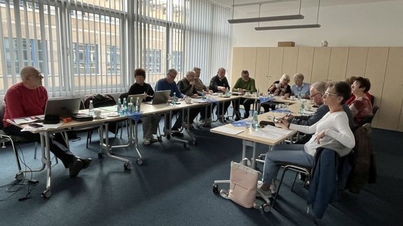 Eine Gruppe von Personen sitzt in einem rechteckigen Besprechungsraum an zusammengestellten Tischen, auf denen Laptops, Unterlagen und Getränkeflaschen stehen. Durch große Fenster fällt Tageslicht in den Raum. Im Hintergrund stehen hohe Schränke, und an der Decke hängt eine längliche Lampe.