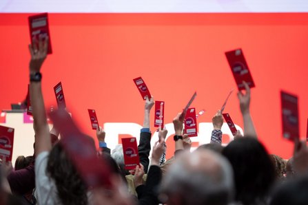 Viele erhobene Arme mit roten Karten vor einer roten Bühnenwand. Die Karten zeigen weiße grafische Elemente und Text. Im unteren Bildbereich sind Köpfe und Schultern von Personen zu sehen, im Hintergrund eine große weiße Beschriftung, teilweise verdeckt.