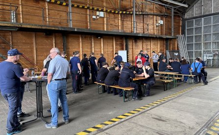 Mehrere Personen sitzen und stehen an Bierzeltgarnituren und Stehtischen in einer großen Halle mit Holzwänden und Betonboden. Einige tragen dunkle Arbeitskleidung. Im Hintergrund sind eine Treppe, Geländer, technische Installationen und ein großes Hallentor sichtbar. Gelb-schwarze Markierungen verlaufen entlang des Bodens.