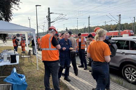 Mehrere Personen in orangefarbenen Warnwesten und Arbeitskleidung stehen neben einem weißen Pavillon auf einem gepflasterten Weg. Links befinden sich Tische mit Getränkebehältern und Geschirr. Im Hintergrund sind zahlreiche Gleise, Oberleitungen und mehrere rote Lokomotiven zu sehen.