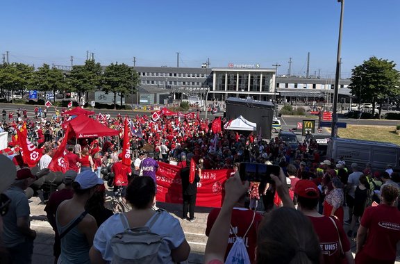 Große Menschenmenge im Freien vor einem Gebäude mit der Beschriftung „DB Hauptbahnhof“. Viele Personen halten rote Fahnen und Banner mit weißem Schriftzug „EVG“. Im Vordergrund stehen weitere Personen auf einer Treppe und schauen auf die Versammlung. Mehrere Zelte, darunter rote Pavillons, sind zwischen der Menge aufgebaut. Rechts stehen ein Fahrzeug und ein Imbissbereich. Blauer Himmel und sonnige Beleuchtung.