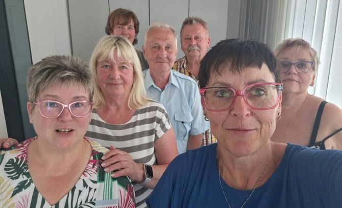 Gruppenfoto in einem Innenraum vor einer hellen Wand mit vertikalen Paneelen. Sieben Personen stehen dicht beieinander und blicken in Richtung Kamera. Im Vordergrund rechts befindet sich eine Person mit kurzer dunkler Frisur, pinkfarbener Brille und blauem Oberteil. Links vorne steht eine Person mit kurzer Frisur, rosafarbener Brille und gemustertem Oberteil. In der Mitte steht eine Person mit blondem Haar und gestreiftem Oberteil. Dahinter sind vier weitere Personen sichtbar, darunter eine Person in hellblauem Hemd, eine Person in kariertem Hemd, eine Person mit Brille sowie eine weitere Person im Hintergrund. Tageslicht fällt von rechts ein.