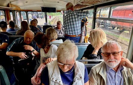 Innenraum eines historischen Triebwagens mit mehreren Sitzreihen. Zahlreiche Personen sitzen auf den Bänken oder stehen im Gang. Durch die großen Fenster sind Gleise, Personen auf einem Bahnsteig und ein roter Güterwagen sichtbar. Über den Sitzen befinden sich Gepäckablagen aus Metallnetz.