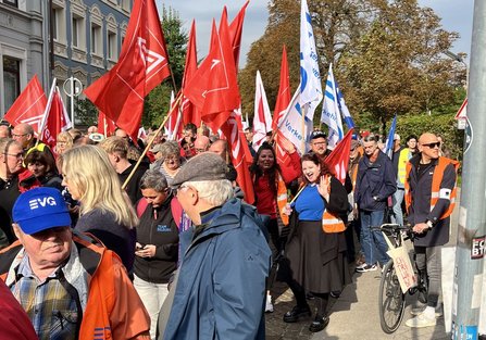 roße Menschenmenge auf einer Straße. Zahlreiche rote und weiße Fahnen mit Logos werden getragen. Mehrere Personen tragen orange Warnwesten, einige blaue Kappen mit der Aufschrift „EVG“. Im Hintergrund stehen mehrstöckige Gebäude und Bäume.