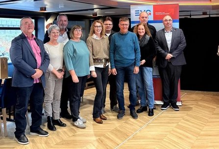 Gruppenfoto in einem Innenraum mit Holzfußboden. Zehn Personen stehen in zwei Reihen vor einem Roll-up-Banner mit dem Schriftzug „EVG“ und einem rot-blauen Hintergrund. Links sind ein Tisch und große Fenster sichtbar. Die Personen tragen überwiegend Freizeit- und Businesskleidung in Blau-, Grau-, Grün-, Schwarz- und Rosatönen. Im Hintergrund befinden sich Deckenleuchten, Fensterflächen und weitere Möbel. Die Aufnahme zeigt die Gruppe von Kopf bis Fuß frontal zur Kamera.