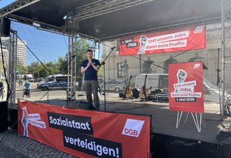 Bühne auf einem Platz mit schwarzem Dachgerüst. Eine Person steht mittig mit einem Mikrofon. Auf der Bühne liegen mehrere Gitarren auf Ständern. Rote Banner mit weißem Text sind angebracht: vorne „Sozialstaat verteidigen!“ mit „DGB“, seitlich ein Plakat „Erst unsere Jobs, dann eure Profite – 1. Mai 2026“. Im Hintergrund sind Gebäude, ein parkendes Fahrzeug und Bäume zu sehen; es ist helles Tageslicht.