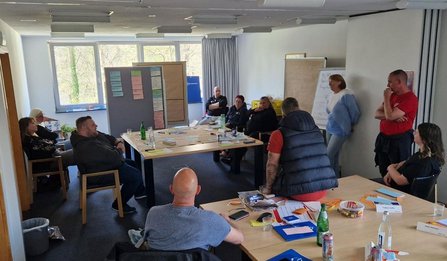 Heller Seminarraum mit mehreren Tischen, die zu einer Arbeitsfläche zusammengeschoben sind. Mehrere Personen sitzen und stehen um die Tische. Auf den Tischen liegen Karten, Papier, Stifte und Flaschen. Im Hintergrund stehen Pinnwände mit angehefteten Karten sowie ein Flipchart; große Fenster lassen Tageslicht herein.
