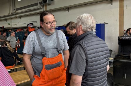 In einer Werkhalle stehen zwei Personen im Vordergrund und unterhalten sich. Eine Person trägt eine orange Latzhose und ein graues T‑Shirt, die andere eine dunkle Steppweste. Im Hintergrund sind weitere Personen, Holzbänke, blaue Spinde, aufgehängte Arbeitskleidung und ein schwarzer Grill zu sehen.
