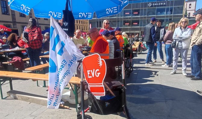 Ansicht eines Platzes mit vielen Menschen zwischen Biertischen. Im Vordergrund steht ein Stuhl mit einem roten Schild mit der Aufschrift „EVG“. Daneben sind Fahnen, darunter eine weiße Fahne mit blauem Logo und Text. Mehrere Personen tragen orangefarbene Warnwesten. Im Hintergrund sind Sonnenschirme, Stände und ein Gebäude mit der Aufschrift „GALERIA“ zu sehen.