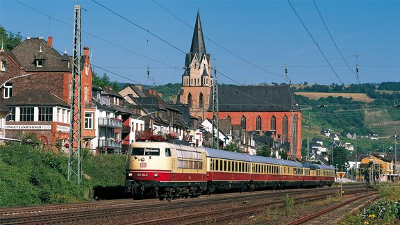 Ein Personenzug mit beigefarbener und roter Lok fährt auf mehrgleisigen Gleisen durch einen Ort mit dicht stehenden Häusern und einer großen Kirche mit hohem Turm im Hintergrund; über den Gleisen spannen sich zahlreiche Oberleitungen, im Hintergrund sind Weinberge und Hügel sichtbar, an den Gebäuden sind Balkone und ein Schild mit der Aufschrift „Weingut Lanius-Knab“ zu erkennen.