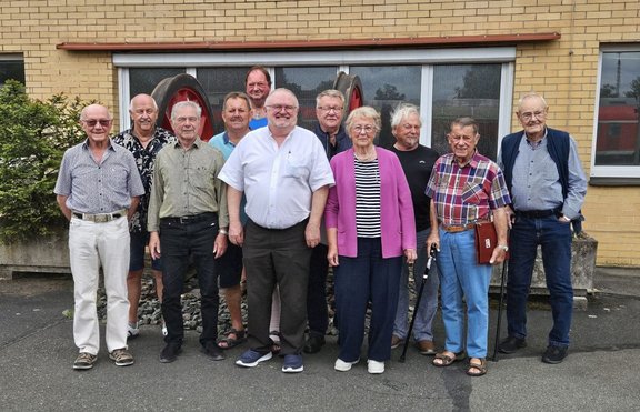 Gruppenfoto von elf Personen vor einem gelben Backsteingebäude. Die Personen stehen in zwei Reihen auf einer befestigten Fläche vor großen Fenstern; im Hintergrund sind zwei große rote Räder sichtbar. Einige Personen tragen Brillen, eine Person hält einen Gehstock. Links steht ein kleiner Nadelbaum, rechts ist durch ein Fenster ein rotes Fahrzeug teilweise zu sehen.
