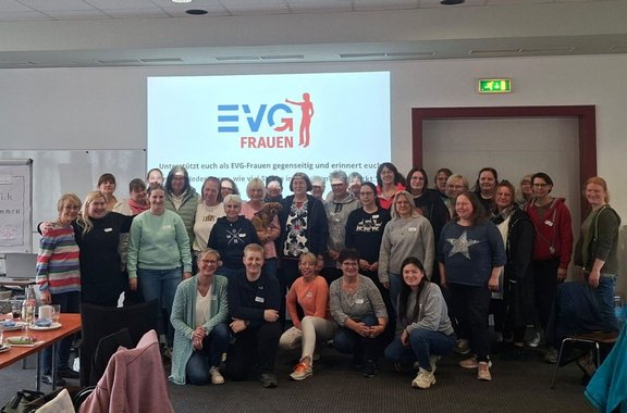 Gruppenfoto in einem Seminarraum mit etwa 30 stehenden und sitzenden Personen. Im Hintergrund ist eine Projektionsfläche mit dem Logo und dem Schriftzug „EVG FRAUEN“ zu sehen; darunter befindet sich mehrzeiliger Text. Der Raum hat weiße Wände, eine abgehängte Decke mit Leuchten und einen Notausgang über einer Tür. Im Vordergrund stehen Tische mit Getränken und Unterlagen sowie Stühle.