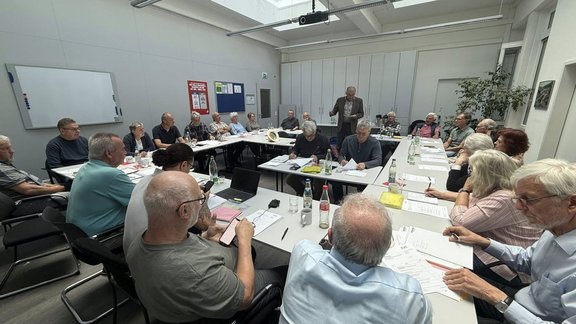 Großer Besprechungsraum mit rechteckigen, zusammengestellten Tischen, an denen viele Personen sitzen. Auf den Tischen liegen Papierunterlagen, Schreibblöcke, Stifte, Laptops, Smartphones, Trinkflaschen, Gläser und Kaffeetassen. Eine Person steht am Kopf der Tischanordnung und hält Unterlagen in der Hand. Die übrigen Personen sitzen rund um die Tische und schauen überwiegend in Richtung der stehenden Person oder auf ihre Unterlagen. An den Wänden befinden sich ein Whiteboard, mehrere Aushänge und Schränke. An der Decke sind Leuchten und ein Beamer angebracht. Im Hintergrund steht eine Pflanze in einem Topf.