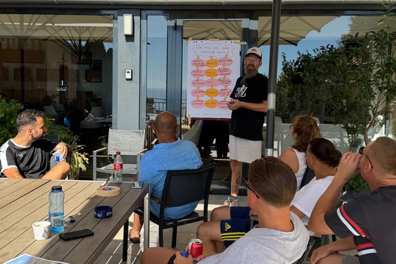 Mehrere Personen sitzen auf einer Terrasse und hören einer Präsentation zu, die an einem Flipchart vorgestellt wird. Im Mittelpunkt steht eine Person vor einem Flipchart mit handschriftlichen Notizen und farbigen Karten. Die Person zeigt auf das Flipchart, während mehrere Teilnehmende auf Stühlen sitzen und in Richtung der Präsentation schauen. Die Gruppe befindet sich an einem großen Holztisch im Außenbereich.
