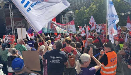 Menschenmenge vor einer Bühne im Freien. Zahlreiche Fahnen und Plakate werden hochgehalten, darunter EVG-Fahnen sowie Schilder mit der Aufschrift „Hart verdient!“. Auf der Bühne ist ein großes Banner mit dem Wort „verdient!“ teilweise sichtbar. Im Vordergrund tragen mehrere Personen Rucksäcke, Kappen und Warnwesten.