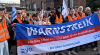 Mehrere Personen in orangefarbenen Warnwesten halten ein großes blau-weiß-rotes Banner. Auf dem Banner steht: „WARNSTREIK“, „Für faire Löhne und Gute Arbeit für alle Beschäftigten!“, „Wir leben Gemeinschaft“ sowie das Logo „EVG“. Daneben sind weiße Fahnen mit EVG-Aufdruck sichtbar. Im Hintergrund stehen Backsteingebäude.