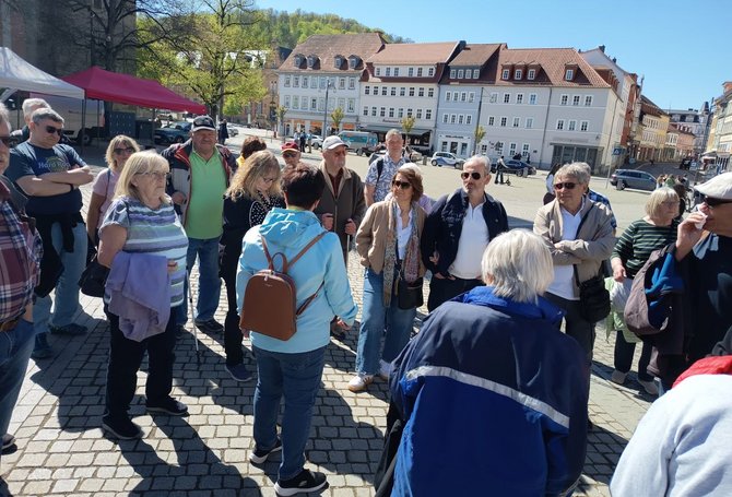 Eine Gruppe von etwa fünfzehn Personen steht auf einem gepflasterten Marktplatz im Freien. Die Personen tragen Freizeitkleidung und Jacken. Im Hintergrund sind mehrstöckige Gebäude mit hellen Fassaden und roten Ziegeldächern zu sehen, außerdem Marktstände mit Zeltdächern. Einige Fahrzeuge stehen am Rand des Platzes, und weitere Personen befinden sich im Hintergrund.