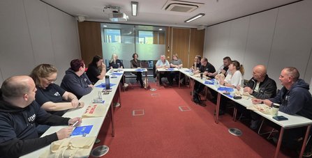 Großer Besprechungsraum mit U-förmig angeordneten Tischen. Mehrere Personen sitzen an den Tischen, auf denen Notizblöcke, Taschen, Trinkflaschen und Papier liegen. An der Stirnseite befinden sich drei weitere Tische mit sitzenden Personen. An der Decke hängen Lampen und ein Beamer; an der Rückwand sind Glasflächen und Holzelemente zu sehen.