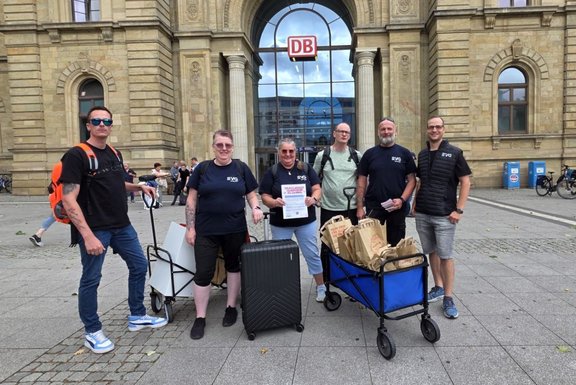 Eine Gruppe von sechs Erwachsenen steht vor dem Eingang eines Bahnhofsgebäudes mit großem „DB“-Logo. Einige tragen dunkle T‑Shirts mit Aufdruck, eine Person hält ein Blatt Papier, andere stehen mit Taschen, Koffer und einem Bollerwagen voller Papiertüten.