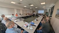 Seminarraum mit U-förmig angeordneten Tischen. Mehrere Personen sitzen an den Tischen und blicken zu einer stehenden Person in der Mitte des Raums. An der Stirnseite ist eine Projektion mit Text auf einer Leinwand sichtbar. Auf den Tischen liegen Unterlagen, Notizblöcke, Smartphones sowie Getränke.