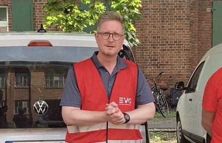 Person in roter EVG-Warnweste und grauem Poloshirt steht vor zwei geparkten Fahrzeugen und einem Backsteingebäude. Die Person trägt eine Brille und hat die Hände vor dem Körper verschränkt. Im Hintergrund sind ein Baum, Fahrräder und weitere Gebäudedetails sichtbar.