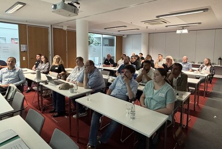 Seminarraum mit mehreren Reihen von Tischen und sitzenden Teilnehmenden. Auf den Tischen stehen Wassergläser, Flaschen, Laptops und persönliche Gegenstände. An der Decke sind Projektoren und Leuchten montiert. Große Fenster und Trennwände befinden sich im Hintergrund.