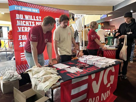 Informationsstand mit rotem Banner. Auf dem Banner steht „EVG JUGEND NRW“ sowie teilweise sichtbarer Text „DU WILLST NICHTS MEHR VERPASSEN?“ und ein QR-Code. Vor dem Tisch stehen drei Personen in roten oder hellen T-Shirts. Auf dem Tisch liegen Stofftaschen, gefaltete weiße Textilien, Schlüsselbänder und Informationsblätter. Rechts gehen weitere Personen an den Ständen vorbei.
